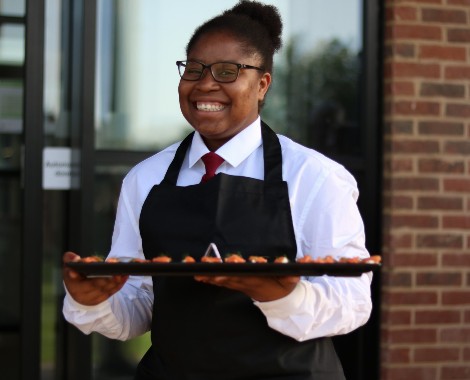 A waitress wearing an apron over a shirt and tie carries a food platter.